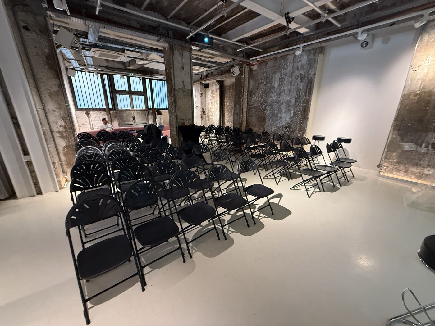 L'ATELIER en configuration conférence : rangs de chaises face à l'écran, vidéoprojecteur au plafond, lumière du jour par la verrière